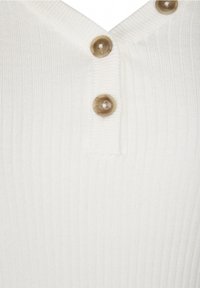 Vitt ribbat tyg med V-halsringning, försett med tre ljusbruna knappar längs fram. Strukturerad yta och rent avslut.