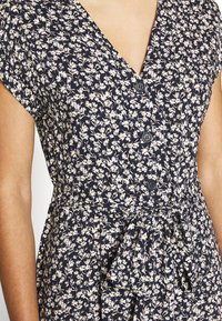 Donna che indossa un vestito nero a maniche corte con una piccola stampa floreale bianca e gialla, con bottoni sul davanti e una cintura annodata in vita.
