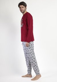 Hombre de pie descalzo, vistiendo una camiseta de manga larga roja y pantalones de pijama grises con patrones de estilo cómic, frente a un fondo blanco liso.