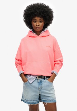 Roze hoodie met een buidelzak, oversized ontwerp en geribbelde manchetten, gedragen over een lichtblauw gestreept shirt en denim shorts.