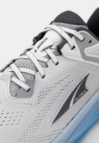 Chaussure de course légère avec une tige en mesh blanc, des accents gris, une semelle bleue contrastante et un branding prominent sur la languette et le côté.