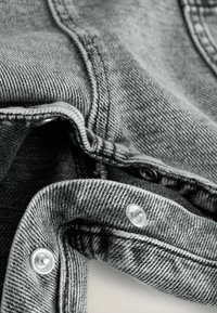 Tela de mezclilla gris con costuras visibles, broches metálicos y una superficie ligeramente texturizada. El primer plano muestra bordes doblados y detalles de botones.