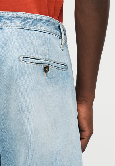 Πλάτη ανοιχτού μπλε τζιν παντελονιού με κουμπωμένη πίσω τσέπη, φορεμένο με κόκκινο πουκάμισο που έχει μπει μέσα και εμφανές σκούρο δέρμα χέρι.