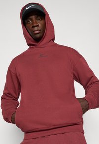 Roter Hoodie mit strukturierter Oberfläche, ausgestattet mit einer Vorder- bzw. Kängurutasche und einem schwarzen Logo. Kombiniert mit einer schwarzen Mütze, die dasselbe Logo zeigt.