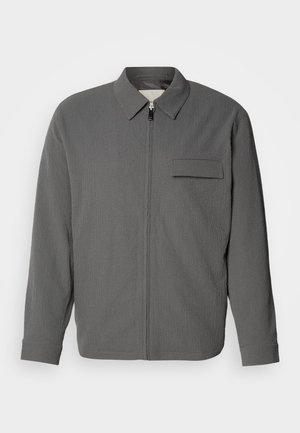 Chaqueta gris con cremallera y tejido texturizado. Cuenta con un cuello tipo polo y un bolsillo en el pecho. Mangas largas con puños.