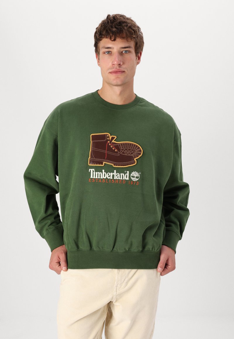 Felpa verde con una grande grafica di un boot marrone e la scritta "Timberland Established 1973". Polsini e vita a coste; texture morbida.