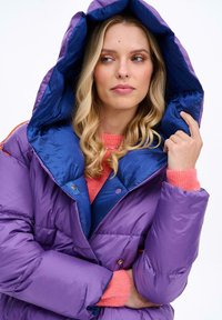 Lesklá fúzička v purpurovej farbe s modrou vnútornou kapucňou. Obsahuje uzáver na gombíky a oversized strih. Modelka má na sebe ružový sveter.