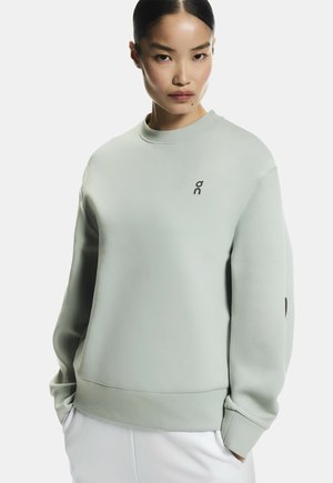 Jeune femme aux cheveux lisses portant un sweat-shirt ample gris clair avec un petit logo noir, mains dans les poches, expression neutre, pantalon blanc.