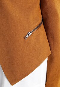 Ruskea kuvioitu takki, jossa on diagonaalinen leikkaus ja hopeinen vetoketju vasemmalla puolella. Materiaali vaikuttaa pehmeältä ja sileältä.