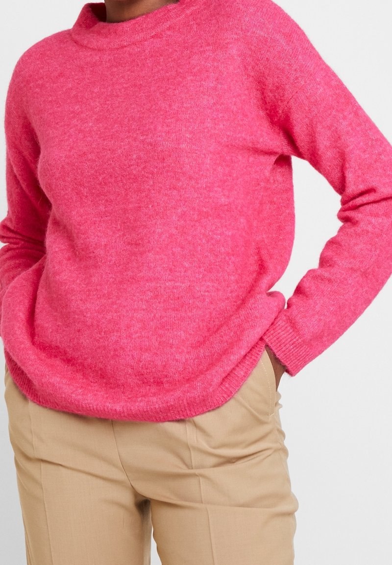 Personne portant un pull duveteux rose vif à manches longues et un pantalon beige, les mains posées négligemment dans les poches, sur un fond uni.