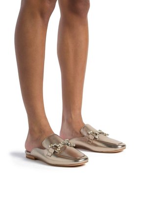 Metallic gold slip-on shoes with a flat sole, featuring a decorative rhinestone-accented strap across the vamp and smooth texture.