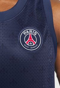 Tmavě modrý sportovní dres bez rukávů s texturovanou, prodyšnou látkou. Obsahuje kulaté logo s nápisy "PARIS" a "SAINT-GERMAIN".