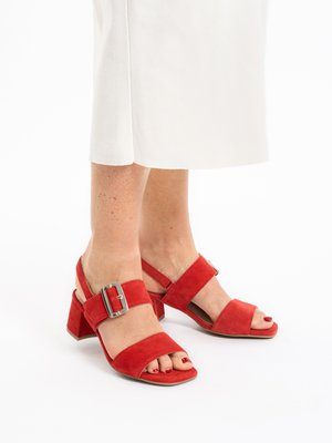 Füße, die rote Sandalen aus Wildleder mit Blockabsatz, Knöchelriemen und silbernen Schnallen tragen, kombiniert mit weißen, knöchellangen Hosen, auf weißem Hintergrund.