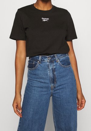 T-shirt noir en coton à manches courtes et col rond, avec un logo Reebok blanc sur la poitrine, associé à un jean bleu taille haute.