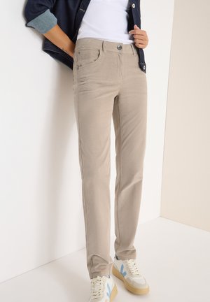 Pantalons beiges en tissu doux, présentant une coupe slim, des poches avant, et une fermeture par bouton, associés à des baskets crème avec des accents bleus.