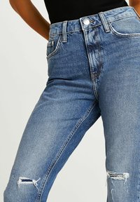 Högmidjade blå denimjeans med slitna knän. Har ljus tvätt, femficksstil och kontrasterande sömmar. Metalldragkedjestängning.