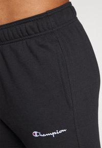 Pantaloni de trening negri, din material elastic, cu talie elastică și buzunar lateral. Au brodat în alb logo-ul "Champion" pe piciorul stâng.