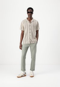 Chemise à rayures à manches courtes en beige et crème, boutonnière sur le devant ; pantalon vert clair ; lunettes de soleil noires ; baskets crème avec semelles marron.
