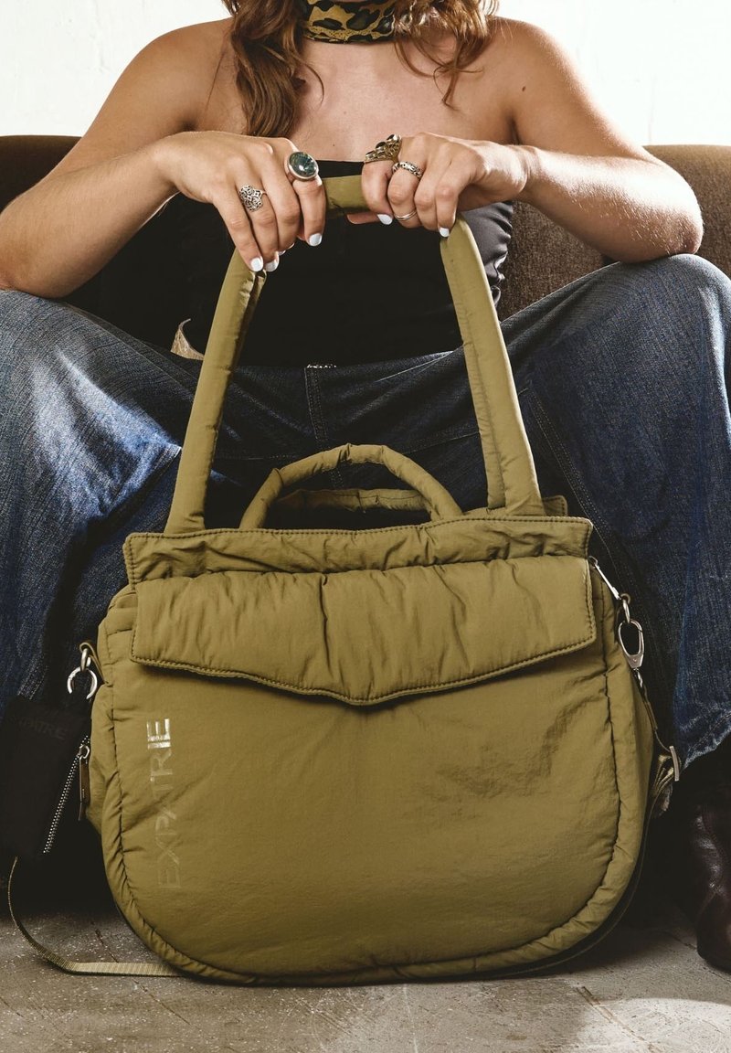 Femme assise en tailleur tenant un sac à main matelassé vert olive avec deux poignées et le mot "EXPERTRE" sur le côté.