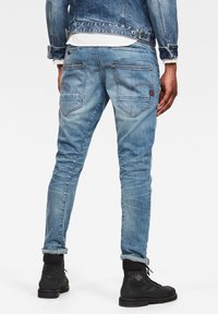 Světle modré džínové kalhoty se slim střihem, pěti kapsami a výrazným prošíváním. Model nosí černé boty.