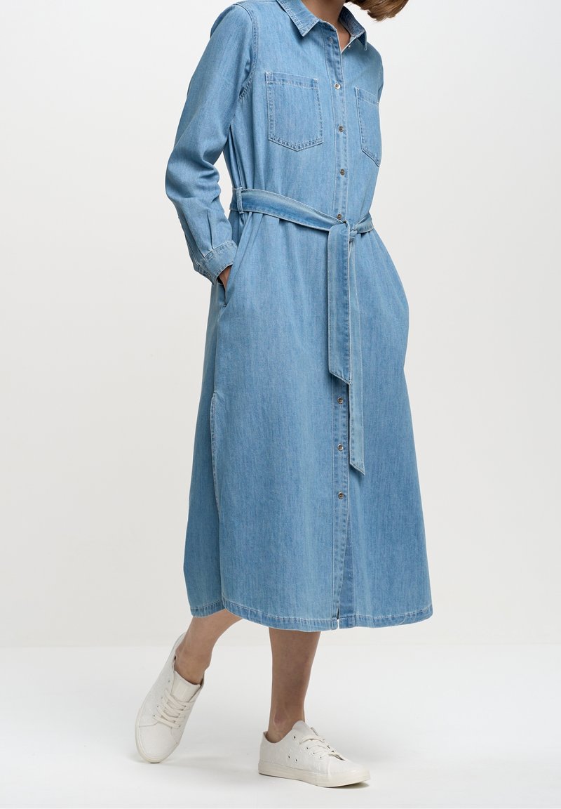 Femme portant une robe en denim bleu clair à manches longues boutonnée, avec des poches poitrine et une ceinture à la taille, associée à des baskets blanches.