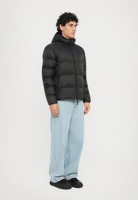 Schwarze Puffyjacke mit Reißverschluss, Kapuze und Logodetail. Das Model trägt hellblaue weit geschnittene Jeans und schwarze Schuhe. Glatte Textur, warmes Design.