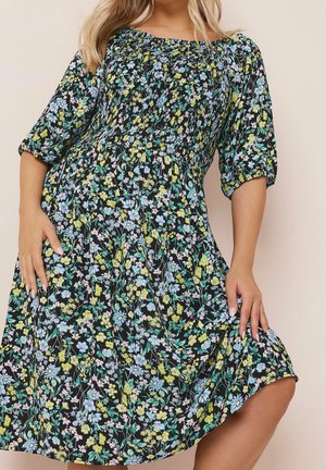 Femme portant une robe midi noire à fleurs avec corsage froncé et manches mi-longues bouffantes, posant avec les mains près de la taille et de l’ourlet.