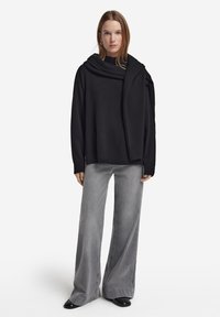 Svart oversized tröja med draperad krage, kombinerad med ljusgrå vida jeans och svarta skor, mot en vit bakgrund.