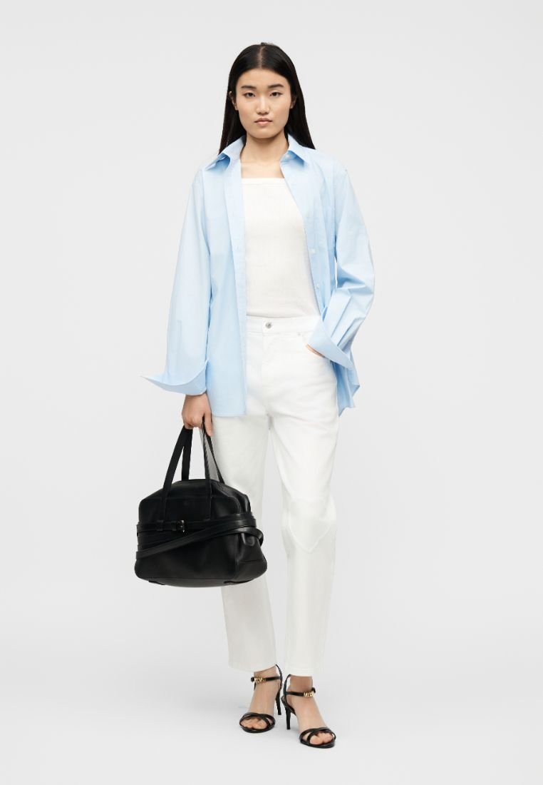 Femme portant une chemise oversize bleu clair, un haut et un pantalon blancs, des sandales à talons noires, tenant un sac à main noir.