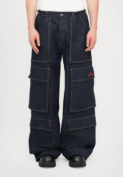 Pantaloni cargo in denim di colore blu scuro, con molte tasche ampie, cuciture gialle a vista e una vita elastica. Scarpe platform nere in basso.
