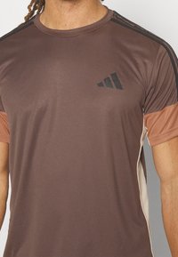 Camiseta deportiva marrón de manga corta, con detalles negros en los hombros y un logotipo en el pecho. Textura suave y corte regular.