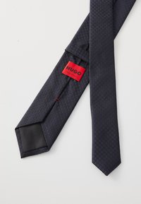 Marineblauer Krawatte mit strukturiertem diagonalen Muster. Sie hat ein schwarzes gestepptes Ende und ein rotes Etikett mit der Aufschrift "HUGO, MADE IN ITALY."