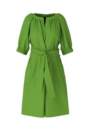 Mujer que lleva un vestido verde brillante hasta la rodilla con mangas hasta el codo y un cinturón atado en la cintura, combinado con sandalias negras de plataforma.