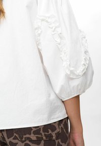 Witte blouse met pofmouwen en franje, gedragen met donkergroene dierenprint pantalon, met een close-up van de arm en taille.