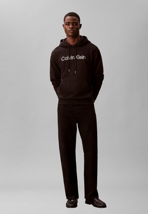 Jeune homme debout portant un sweat à capuche noir Calvin Klein, un pantalon noir et des chaussures noires sur un fond gris clair uni.