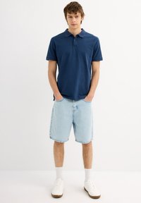 Camiseta polo azul marino de manga corta, combinada con shorts de mezclilla claros hasta la rodilla. Zapatillas blancas con suelas marrones y calcetines blancos.