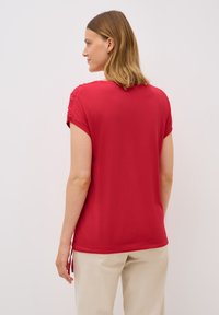T-shirt léger rouge à manches courtes, avec des détails froncés aux épaules et une coupe décontractée, associé à un pantalon neutre.