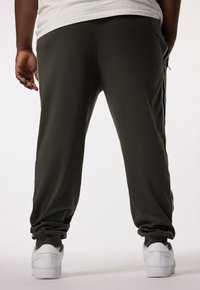 Olijfgroene sweatpants met een ontspannen fit, voorzien van zwarte zijstrepen en elastische boorden. Draagt witte sneakers, weergegeven van achteren.