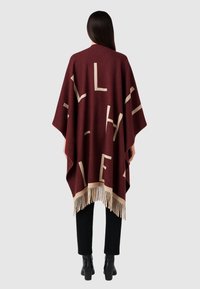 Poncho bordeaux con grandi lettere beige, orlo frangiato, texture morbida e vestibilità ampia, indossato sopra pantaloni neri e stivali alla caviglia.