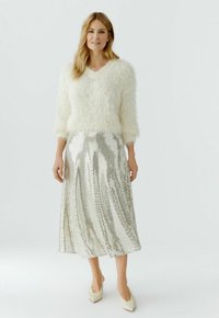 Vrouw die een crèmekleurige, pluizige trui draagt, een zilveren pailletten midirok en crèmekleurige pumps met een puntige neus, staande tegen een witte achtergrond.