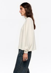 Kremowy dzianinowy sweter z wysokim kołnierzem, luźnym fasonem i prążkowanymi mankietami, zestawiony z ciemnoniebieskimi dżinsami. Miękka faktura, minimalistyczny design.