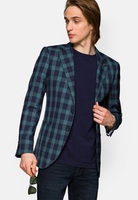 Skrzyżowany marynarka w ciemnozielonym i granatowym materiale. Posiada dwa guziki, klapy w kształcie litery notch oraz jedną kieszeń na piersi. Nosi się ją na granatowej koszulce.