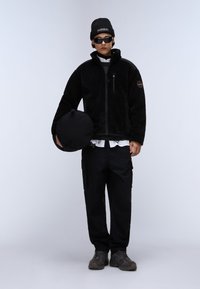 Μαύρο φλις μπουφάν με φερμουάρ, πλαϊνές τσέπες και λογότυπο, συνδυασμένο με μαύρα cargo παντελόνια και γκρι αθλητικά παπούτσια. Το μοντέλο κρατάει μια στρογγυλή μαύρη τσάντα.