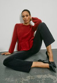 Donna con maglione rosso, pantaloni neri e tacchi neri seduta a terra contro una parete bianca, che posa con una mano dietro la testa.