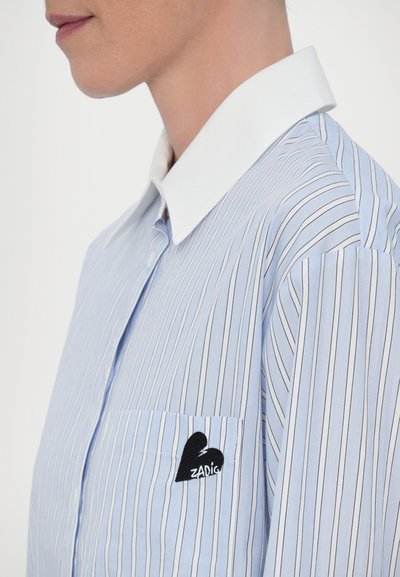 Light blue striped shirt with a white collar, featuring a left chest pocket adorned with a black heart logo reading "ZADIG."