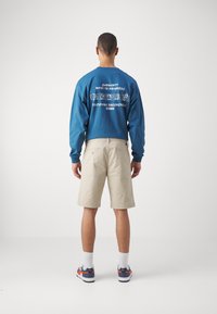 Blaues Langarmhemd mit weißem Text und Grafik auf dem Rücken, kombiniert mit beigen Shorts. Model trägt weiße Socken und blaue Sneakers mit orangefarbenen Akzenten.