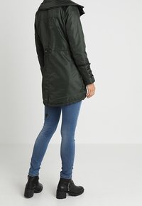 Parka imbottita verde con cappuccio, vita con coulisse e dettagli neri. Abbinata a jeans skinny blu e stivaletti neri.