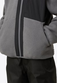 Siva flis jopica z zgornjim delom iz črnega najlona, ki ima sprednji zadrgo in stranske žepe. Gladek teksturni material z kontrastnim oblikovalskim elementom.