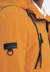 Veste imperméable orange avec fermeture éclair sur le devant, capuche ajustable et patch de marque noir sur la manche. Présente un tissu texturé et des détails fonctionnels.