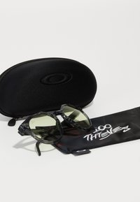 Sonnenbrille mit einem schwarz-grau marmorierten Rahmen, dunklen grünen Gläsern, begleitet von einem schwarzen Etui und einem Mikrofasertäschchen mit Logo.